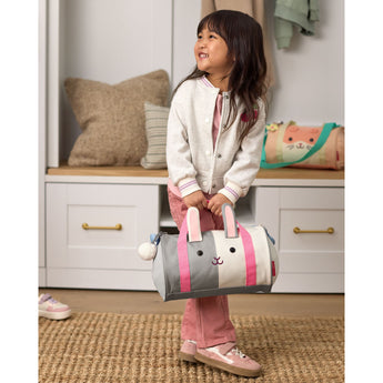 Smiling child holds a gray and white Skip Hop Duffimals bunny duffle bag with pink ears and handles.