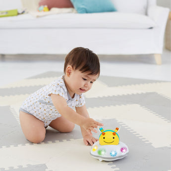 Baby in a white onesie reaching for a Skip Hop Explore & More Follow Bee Crawl Toy with colorful lights.