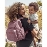 Smiling mom carries a baby while wearing a dusty rose quilted Skip Hop Forma backpack.