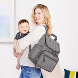 Smiling mom holds baby, wearing a gray quilted Skip Hop Forma backpack nappy bag with side pockets.