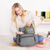 Smiling woman packing a gray quilted Skip Hop Forma backpack with a mesh packing cube.