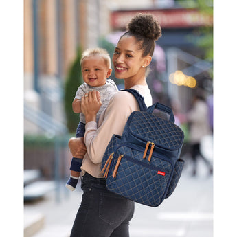 Smiling woman holds baby, wearing a navy quilted Skip Hop Forma Backpack with tan accents.
