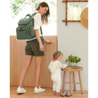 Woman wearing a green quilted Skip Hop Forma backpack, standing by a door with a baby.