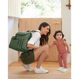 Woman wearing a green quilted Skip Hop Forma backpack, crouching next to a baby in a home entryway.