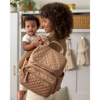 Woman carrying baby and a stylish tan quilted Skip Hop Forma backpack nappy bag with gold zippers.