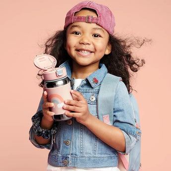 Smiling child holding a pink Skip Hop stainless steel straw water bottle with a rainbow design.