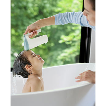 Mom uses a white and light blue Skip Hop Moby Waterfall Rinser to rinse a happy child's hair in a white bathtub.