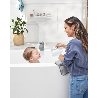 Smiling woman rinses baby's hair with a white Skip Hop Moby Waterfall Rinser in a white bathtub.
