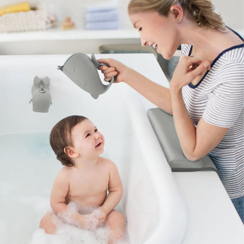 Happy baby in a tub, looking at a mom holding a gray whale-shaped Skip Hop Moby Waterfall Rinser.
