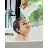 Hand holding a white and blue Skip Hop Moby Waterfall Rinser over a smiling child's head in a white tub.