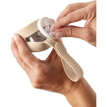 Hands placing a white pacifier into a beige Skip Hop silicone pacifier holder with a strap.