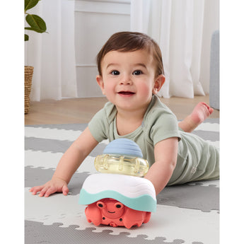 Happy baby crawling on a gray and white mat, playing with a colorful Skip Hop Stack & Crawl 4-in-1 Crab toy.