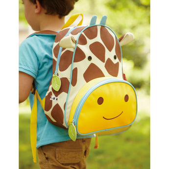 Toddler wearing a Skip Hop Zoo giraffe backpack with brown spots, yellow accents, and a smiling front pocket.