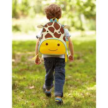 Toddler boy wearing a Skip Hop Zoo Giraffe Backpack with brown spots and yellow face, walking in grass.