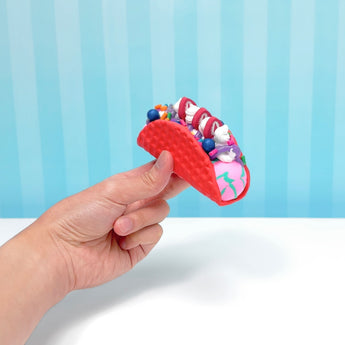 Hand holding a red slime taco filled with colorful, swirled slime, sprinkles, and blueberries against a blue striped background.