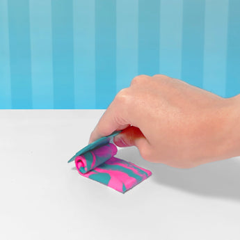 Hand rolling vibrant pink and teal swirled slime with a blue tool on a white surface, blue striped background.