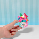 Hand holds a small pink and teal cup filled with colorful slime resembling ice cream, topped with candy and a green leaf.