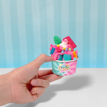 Hand holds a small pink and teal cup filled with colorful slime resembling ice cream, topped with candy and a green leaf.