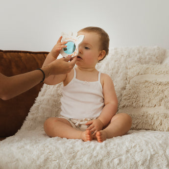 Baby in white tank top holding a Sophie la girafe Magic Mirror SIGHT White teething toy.