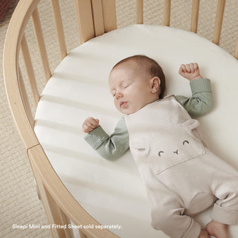 Stokke Sleepi Mini white polyurethane foam mattress pad in a wooden crib with a sleeping baby.
