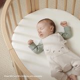 Baby sleeping soundly on a white Stokke Sleepi Mini polyurethane foam mattress in a natural wood crib.