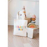 White tote bag with colorful text 'DELANEY SHAY' next to wooden toys on a light wooden floor.